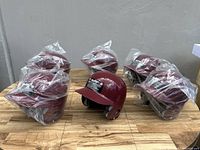 Photo showing six Rawlings maroon batting helmets, five wrapped in plastic and one unpackaged, placed on a wooden surface.