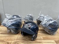 Photo showing six navy blue Rawlings batting helmets, five wrapped in plastic and one unwrapped, placed on a wooden surface.
