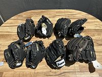 Eight baseball gloves laid out on wood surface, varying black and dark brown leather with white lace details and tags visible.