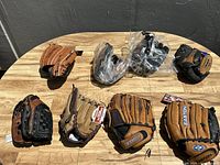 Photo of eight baseball gloves arranged on a wooden surface, showing a variety of colors and brands including Easton, some new with tags, some used.