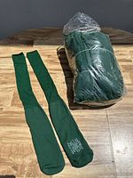 Pair of hunter green sports socks laid flat beside a large sealed plastic bag containing multiple more pairs of the same socks.