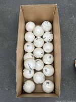 Top-down view of a cardboard box containing approximately 18 Dudley brand softballs with visible barcodes.