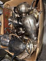 Box of assorted silver plate tea pots, pitchers, and other vessels showing tarnished surfaces and dark handles.