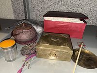 Overview photo of brass jewelry box, reddish-brown teapot, glass jar with orange lid, various jewelry pieces, and red box with tissue paper on shelf.