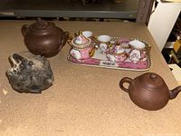 Group of teapots including Yixing clay, carved wooden turtle teapot, and miniature pink porcelain tea set on a tray, displayed on a shelf.