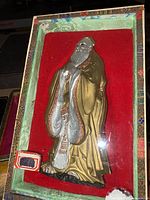 Brass-colored figurine of elderly man in flowing robes shown inside a decorative box with red velvet lining.