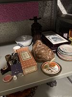 Table showing a collection of miscellaneous items: ceramic plates, bowl, Buddha figurine, Asian-style bottle, framed artwork, pipes, and small containers.