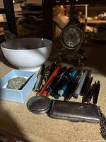 Overview showing assorted pens, small ornate clock, white bowl with items, small blue box with silver jewelry, pocket watch, and silver case on chain.