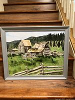 Framed oil painting of rustic farm buildings in green countryside with hills and trees.