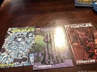 Three Teenage Mutant Ninja Turtles comics with distinct Comic-Con themed covers placed on wooden surface.
