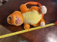 Front side view of the Charmander plush backpack with an orange body, beige belly, and embroidered facial details, lying on table with measuring tape showing length approximately 30cm.