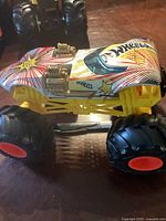 Side angled view of a large Hot Wheels monster truck showing white body with colorful starburst graphics, yellow chassis, black large wheels with red rims, and gold engine details.