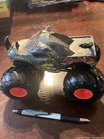 Side view of large monster truck toy with rhino themed design, showing detailed textured body and large black wheels with red centers, next to pen for scale.