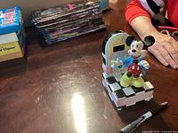 Front angle showing Mickey Mouse figure, jukebox design, and base on wood table with surrounding items.