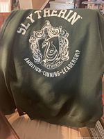 Back view of dark green hoodie showing large white Slytherin crest design with serpent and words 'Slytherin Ambition Cunning Leadership'.