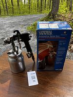 Spray gun with attached metal fluid canister next to its box on wooden surface.