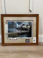 Framed commemorative print titled "Lifting to the North" by Ken Zylla showing ducks flying over a lake, framed in light oak with linen matting and inset stamp.