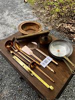Full lot of copper kitchen utensils and cookware on wooden table outdoors showing ladles, spatulas, kettle, basin, frying pans, and brass rod