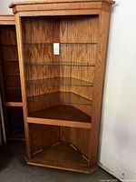 Full frontal view of the corner cabinet showing the oak wood frame and four glass corner shelves inside.