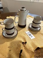 Photo showing the Denby coffee pot with lid, two stacks of coffee cups, and saucers on a wooden surface.