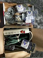 Top-down view of two cardboard boxes containing electrical items including mounted metal boxes, packaged mechanical dial timers, power strips, and cords.