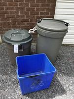Three used waste management containers on the ground: large gray Rubbermaid Animal Stopper trash can, smaller gray trash can with a lid, blue recycling bin with recycling symbol.