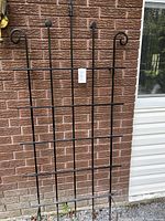 Full front view of the black iron garden trellis leaning against a brick wall and vinyl siding.