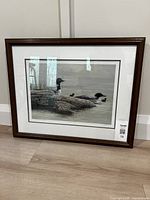 Framed print of a loon family on water and rocks, showing the full artwork, matting, and dark wooden frame.