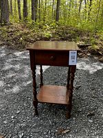 Full view of the small solid wood table showing drawer, turned legs, and lower shelf, placed outdoors on gravel with forest background.