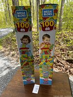 Two boxes of Mega Bloks First Builders 100 piece building blocks standing upright outdoors on a wooden surface, showing front and side of packaging with colorful block graphics.