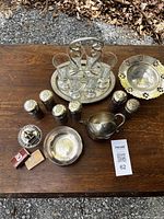Full view of all silver items including three sets of salt and pepper shakers, tray with six liquor glasses, match holders, and bowls on wooden surface.