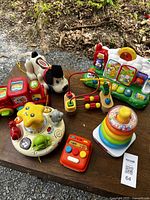 Six toddler toys including a pull-along dog, musical keyboard, star-shaped activity toy, stacking rings, and small red electronic toy arranged on a wooden surface outdoors.