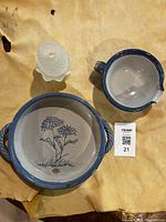Photo showing three items: a round pottery dish with blue handles and blue floral design inside, a smaller pouring pottery dish with blue rim, and a clear glass candle holder with textured surface.