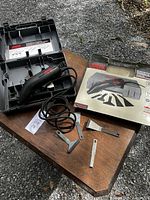 Electric SKIL scraper tool with power cord, three metal attachments laid out, carrying case open on wooden surface.