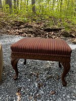 Photo of stool showing the striped cushioned upholstery and carved wooden legs outdoors on gravel