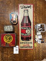 Photo showing all items including the Nichol Kola sign, three tins, a glass jar with vintage matchbooks spread beside it, on a wood surface.