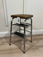 Full view of the step stool showing chrome metal frame, two steps, and brown padded seat in the down position.