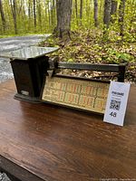 Front perspective of vintage black postage scale on a wooden surface outdoors showing the flat metal weighing platform and colorful postal rate chart.