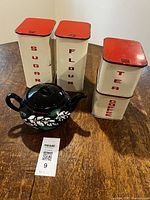 All canisters with red sliding tops and black ceramic teapot on wooden table