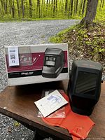 Welding mask with box, gloves, and manual on a wooden table outdoors.