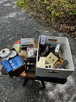 Wide view of the workshop lot showing box filled with electrical switch plates, tools, hardware and wiring items on a bench outdoors.