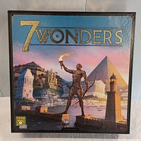 Front cover of the 7 Wonders board game box showing artwork of ancient wonders and a statue holding a torch.