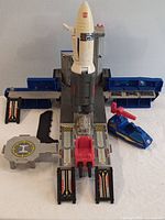 Full view of vintage Transformers Countdown base including white rocket, grey launchpad with blue accordion style extending platforms and red accents. Blue turret vehicle positioned beside base.