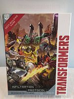 Front box cover of Transformers Infiltration Protocol expansion deck building game showing robot characters and logo.