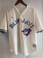 Front view of white baseball jersey with 'BLUE JAYS' text and team logo, hanging on hanger.