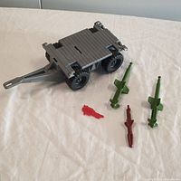 Gray toy trailer with four black wheels and tow hitch shown with green missile launchers, brown missile, and translucent red gun on white surface.