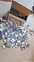 Open white cardboard box with numerous Magic: The Gathering cards scattered on a beige carpet. Cards include various colors and characters, some sleeved.