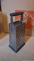 Front angled view of four-sided metal cheese grater showing medium and small grating holes with black plastic handle and rubber base.