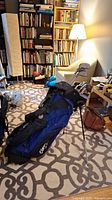 Blue Cleveland golf bag standing on patterned carpet in living room with bookshelves in background, showing full set of golf clubs inside.