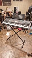 Black Concertmate 980 electronic keyboard on black adjustable X-style metal stand with music rest, in a room with various items and a tile floor.